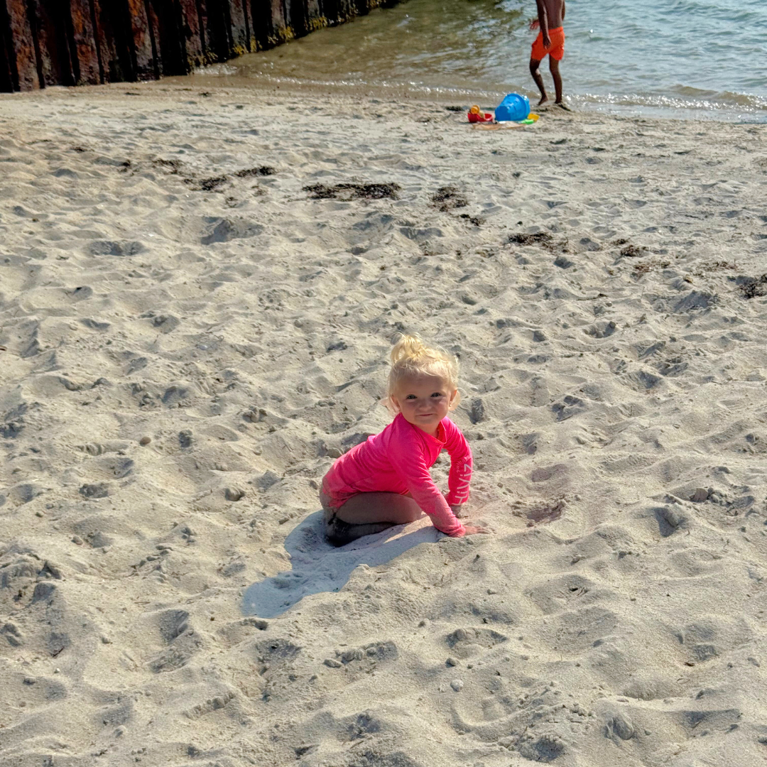 Child in neon pink rash guard playing in sand on a beach with water and another person in the background in neon orange swim trunks